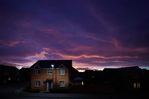 This Urban landscape photograph shows a residential street in Chesterfield during an evening sunset in winter. The main subject of the image is a suburban house illuminated by a streetlight, with other houses visible in the background. The sky is filled with dramatic hues of purple and orange, showcasing nature’s vibrant colors as the sun sets behind the rooftops. The photograph captures the tranquil transition between day and night, highlighting the interplay between the built environment and the natural beauty of the sunset sky in this urban setting.